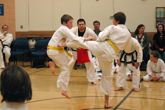Two yellow belt boys spar at a tournament. The one standing lands a kick to the mid-section of the one jumping in.