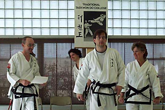 Three men and a woman, all in uniforms, smiling with a poster of Bailey's Traditional Taekwon Do College in the  background.